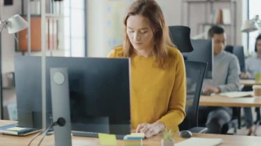 Güzel ve zeki kızıl saçlı kadın uzmanı masasında oturuyor. Masaüstü bilgisayarında çalışıyor. Yetenekli Yaratıcı İnsanlar İçin Ergonomik Mobilyalı Parlak ve Modern Açık Uzay Ofisi