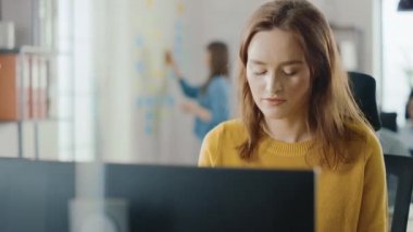 Güzel ve zeki kızıl saçlı kadın uzmanı masasında oturuyor. Masaüstü bilgisayarında çalışıyor. Yetenekli Yaratıcı İnsanlar İçin Ergonomik Mobilyalı Parlak ve Modern Açık Uzay Ofisi