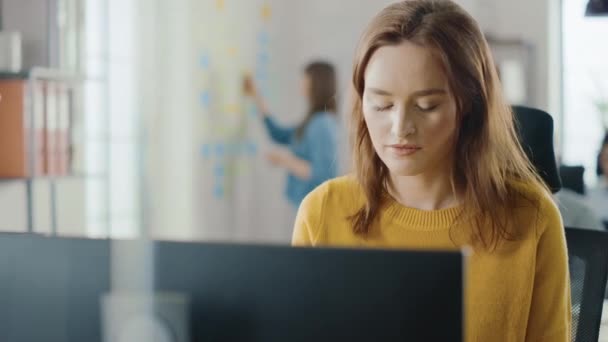 美しくスマートな赤髪の女性専門家は、デスクトップコンピュータ上の彼女の机の仕事に座っています。才能創造的な人々のためのスタイリッシュな人間工学に基づいた家具を備えた明るくモダンなオープンスペースオフィス — ストック動画