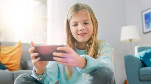 Портретний знімок розумної милої дівчини, що сидить на килимі, грає у відеогрі на своєму смартфоні, тримає і використовує мобільний телефон в горизонтальному ландшафтному режимі. Дитина весело грає відеоігри в сонячній вітальні . — стокове фото