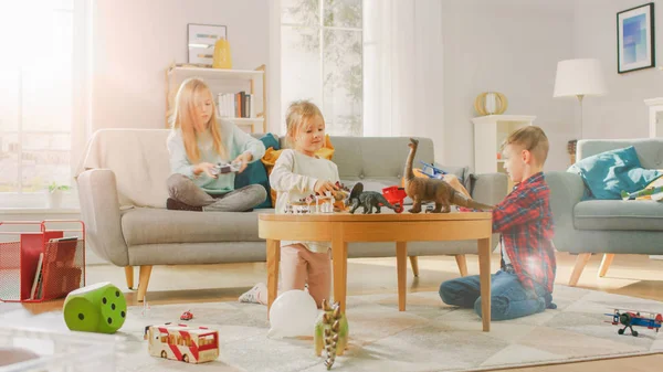 Em casa: Menina bonito jogando no console de videogame, Usando Joystick Controller, Seu Brothe mais jovem joga com avião de brinquedo e irmã mais nova joga com dinossauros de brinquedo. Crianças felizes brincando na sala de estar ensolarada . — Fotografia de Stock