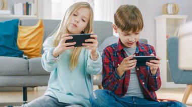 Evde Bir Halı üzerinde Oturan: Sevimli Küçük Kız ve Sweet Boy İki Akıllı Telefonlar üzerinde Rekabetçi Video Oyunu Oynamak, Yatay Manzara Modunda onları tutarak.