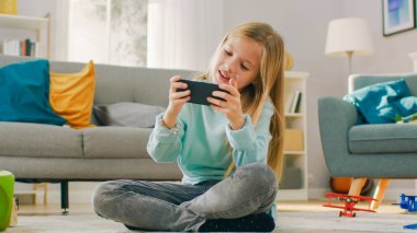 Akıllı Sevimli Kız Onun Smartphone Video Oyunu Oynarken Evde Halı Üzerinde Oturan, Tutar ve Yatay Manzara Modunda Cep Telefonu kullanır. Çocuk Güneşli Oturma Odasında VideoOyunu Oynamaktan Eğlenceli.