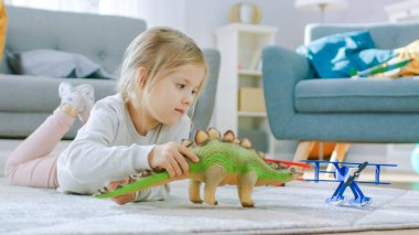 Sevimli Küçük Kız Sarışın Evde Bir Halı üzerinde döşeme, Oyuncak Dinozorlar ve Uçaklar ile çalış. Sunny Living Room Oyuncaklar ile Oynayan Mutlu Çocuk. Yakın Çekim Portre Çekimi.