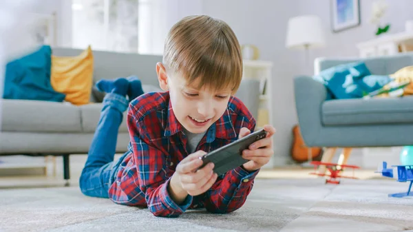Smart Little Boy Laying on a Carpet Plays in Video Game on His Smartphone, Holds Mobile Phone in Horizontal Landscape Mode. Child Has Fun Playing Videogame in Sunny Living Room.