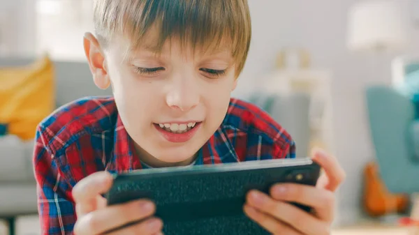 Close-up portret van een slimme kleine jongen leggen op een tapijt speelt in video game op zijn smartphone, houdt mobiele telefoon in horizontale landschap modus. Kind heeft plezier spelen video game in zonnige woonkamer. — Stockfoto