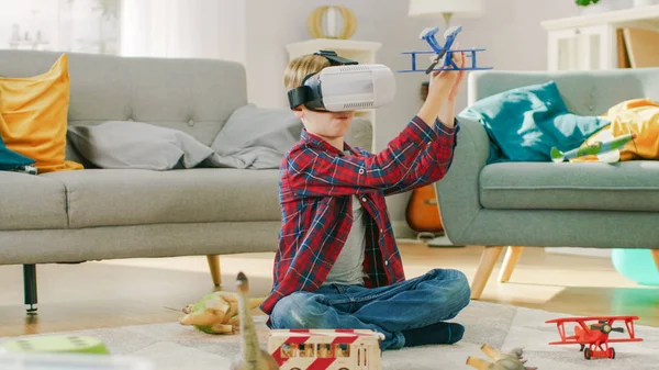 Smart Little Boy Wearing Virtual Reality Headset Plays with Toy Airplane while Sitting on a Carpet in His Living Room. Happy Child Uses Futuristic AR Glasses at Home.