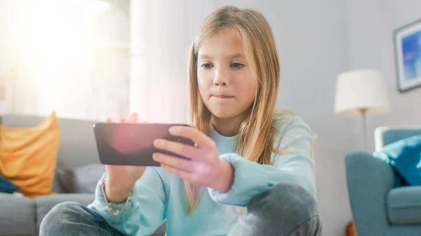 Retrato de una chica linda e inteligente sentada en una alfombra jugando en un videojuego en su teléfono inteligente, sostiene y utiliza el teléfono móvil en modo paisaje horizontal. Niño se divierte jugando videojuego en la sala de estar soleada . — Foto de Stock