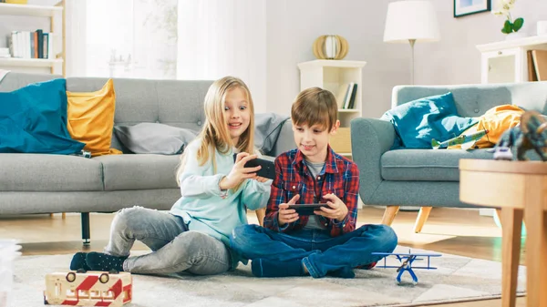 At Home Sitting on a Carpet: Cute Little Girl and Sweet Boy Playing in Competitive Video Game on two Smartphones, Holding them in Horizontal Landscape Mode. Menina ganha e celebra . — Fotografia de Stock