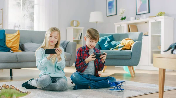 En casa sentado en una alfombra: Linda niña y dulce niño jugando en el videojuego competitivo en dos teléfonos inteligentes, sosteniéndolos en modo paisaje horizontal . — Foto de Stock