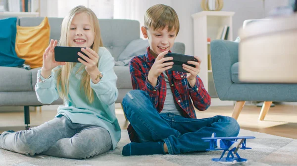 A casa seduti su un tappeto: carina bambina e dolce ragazzo che giocano in un videogioco competitivo su due smartphone, tenendoli in modalità orizzontale paesaggio . — Foto Stock