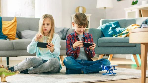 Em casa sentado em um tapete: Menina bonito e doce menino jogando em vídeo game competitivo em dois smartphones, segurando-os no modo paisagem horizontal . — Fotografia de Stock