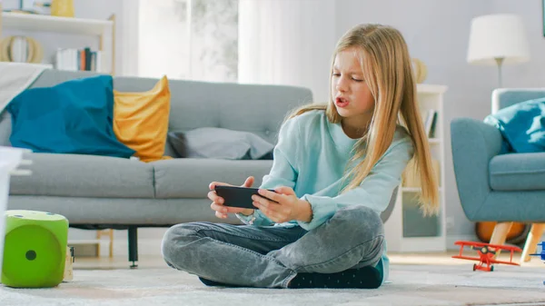 Smart cute girl zittend op een tapijt thuis spelen in video game op zijn smartphone, houdt en gebruikt mobiele telefoon in horizontale landschap modus. Kind heeft plezier spelen video game in zonnige woonkamer. — Stockfoto
