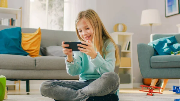 Smart cute girl zittend op een tapijt thuis spelen in video game op zijn smartphone, houdt en gebruikt mobiele telefoon in horizontale landschap modus. Kind heeft plezier spelen video game in zonnige woonkamer. — Stockfoto