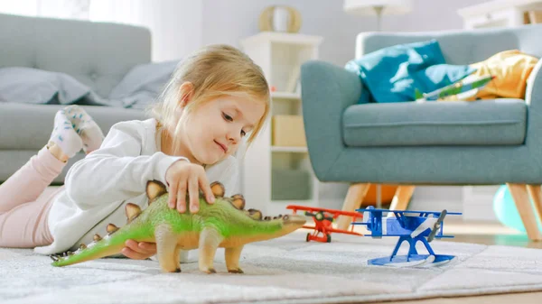 Schattig klein meisje blond leggen op een tapijt thuis, speelt met speelgoed dinosaurussen en vliegtuigen. Gelukkig kind spelen met speelgoed in zonnige woonkamer. Close-up portret foto. — Stockfoto