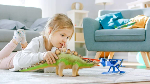 Schattig klein meisje blond leggen op een tapijt thuis, speelt met speelgoed dinosaurussen en vliegtuigen. Gelukkig kind spelen met speelgoed in zonnige woonkamer. — Stockfoto