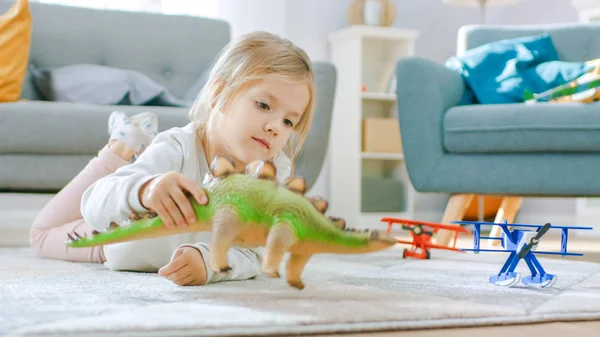 Søt, liten blond jente som ligger på et teppe hjemme, spiller med Toy Dinosaurer og fly. Happy Child leker med leker i solfylte stue. Avgrenset portrett Skudd . – stockfoto