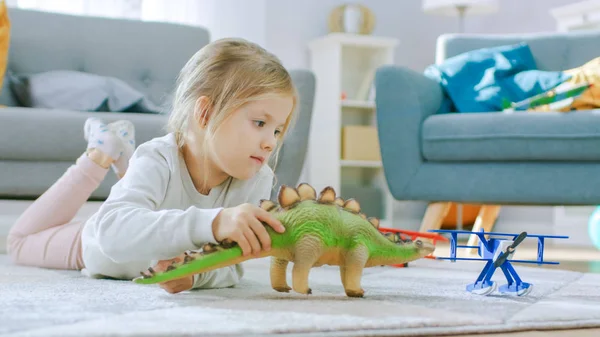 Schattig klein meisje blond leggen op een tapijt thuis, speelt met speelgoed dinosaurussen en vliegtuigen. Gelukkig kind spelen met speelgoed in zonnige woonkamer. Close-up portret foto. — Stockfoto