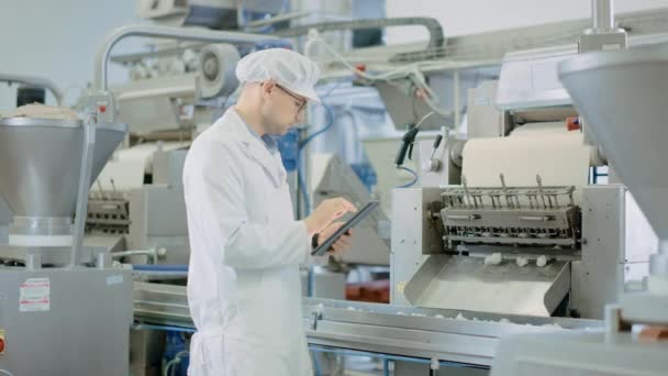 Young Male Quality Supervisor or Food Technician está inspeccionando la producción automatizada en una fábrica de Dumpling Food. Empleado Utiliza una Tablet Computer for Work. Lleva un sombrero sanitario blanco y una bata de trabajo . — Vídeo de stock