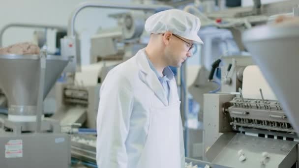 Young Male Quality Supervisor or Technician in Glasses está trabajando en una fábrica de alimentos. El empleado mira y sonríe en la cámara. Expresa Éxito y Felicidad. Lleva un sombrero sanitario blanco y una bata de trabajo . — Vídeo de stock