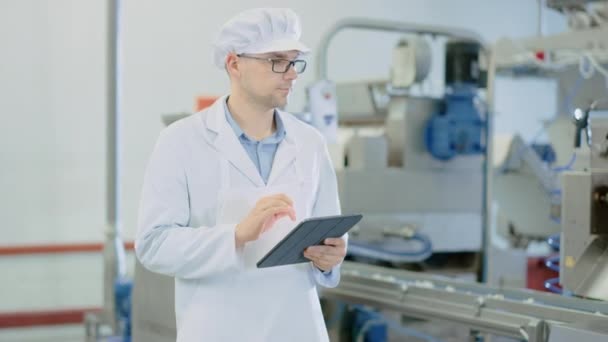 若い男性品質監督者または食品技術者は、餃子食品工場で自動生産を検査しています。従業員は、作業にタブレット コンピュータを使用します。彼は白い衛生帽と仕事のローブを着ている. — ストック動画
