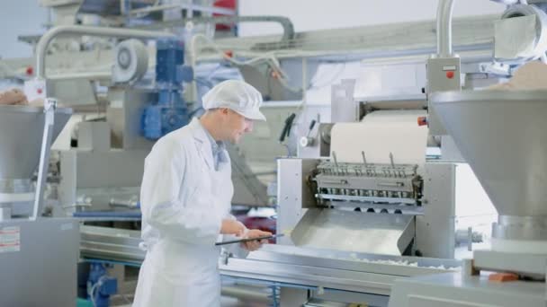 Ο επόπτης ποιότητας νεαρής ηλικίας ή ο τεχνικός τροφίμων επιθεωρούν την αυτοματοποιημένη παραγωγή σε ένα εργοστάσιο φαγητών ζυμαρικών. Ο εργαζόμενος χρησιμοποιεί έναν υπολογιστή tablet για εργασία. Κοιτάζει την κάμερα και τα χαμόγελα. — Αρχείο Βίντεο