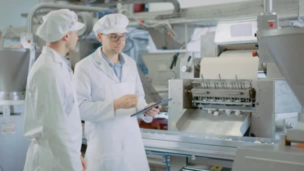 İki Genç Erkek Kalite Yöneticisi veya Gıda Teknisyeni, Bir Hamur köftesi fabrikasında Otomatik Üretimi inceliyor. Çalışan, çalışmak için bir tablet bilgisayar kullanır. Beyaz iş elbisesi giyiyorlar.. — Stok video