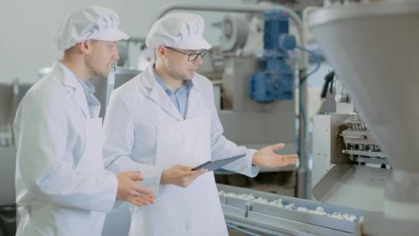 Dos jóvenes supervisores de calidad masculina o técnicos de alimentos están inspeccionando la producción automatizada en una fábrica de alimentos de Dumpling. Empleado Utiliza una Tablet Computer for Work. Llevan ropas blancas de trabajo. — Vídeo de stock