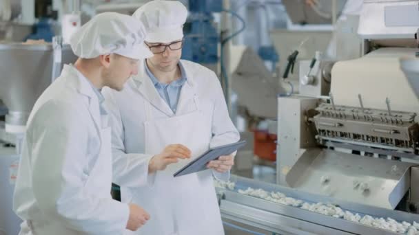 Two Young Male Quality Supervisors or Food Technicians are Inspecting the Automated Production at a Dumpling Food Factory. Employee Uses a Tablet Computer for Work. They Wear White Work Robes. — Stock Video