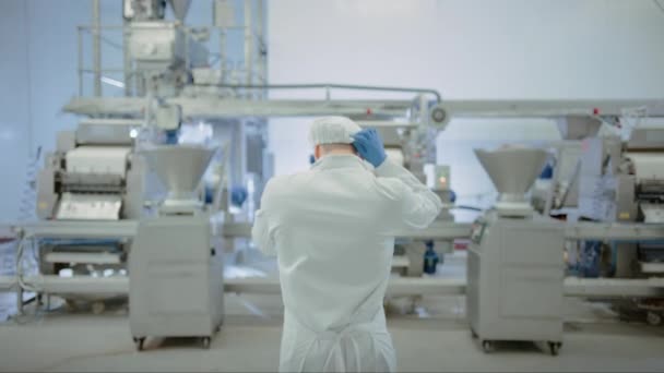餃子工場の若い男性食品従業員がコンベアベルトに来ます。彼は同僚と一緒に働き始める。白い衛生用帽子とラテックス手袋を着用する。. — ストック動画
