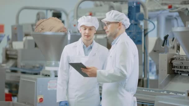 Zwei Mitarbeiter der Young Food Factory diskutieren über arbeitsbezogene Themen. Männliche Techniker oder Qualitätsmanager verwenden einen Tablet-Computer für die Arbeit. Sie tragen weiße Hygienemütze und Arbeitskleidung. — Stockvideo