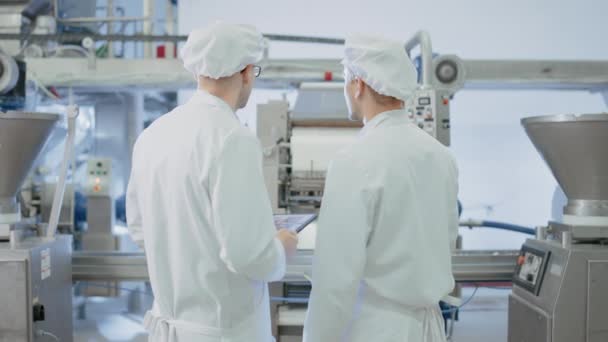 Dos jóvenes empleados de la fábrica de alimentos discuten asuntos relacionados con el trabajo. El Técnico Masculino o Gerente de Calidad utiliza una Tablet Computer para el Trabajo. Llevan sombrero sanitario blanco y ropas de trabajo. — Vídeo de stock