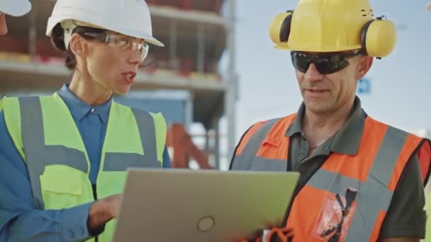 Une équipe diversifiée de spécialistes utilise un ordinateur portable sur le chantier de construction. Projet de construction immobilière avec des machines : Ingénieur civil, investisseur, femme d'affaires et constructeur discutant du plan directeur — Video