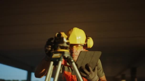 Dentro del sitio de construcción de edificios comerciales / industriales: el ingeniero profesional Surveyor toma medidas con teodolito, usando Tablet Computer digital. En el fondo Rascacielos Marcos de encofrado y grúa — Vídeo de stock