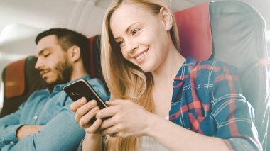 Yönetim Kurulu, ticari uçak güzel genç sarışın kullanır smartphone cep telefonu ile onun İspanyol ise erkek komşu uyur. Uçak pencereden güneş parlıyor.