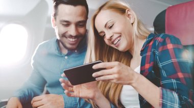 Ticari uçak bir gemide güzel genç sarışın yakışıklı İspanyol erkek ile kullanma Smartphone ve gülmek. Uçak pencereden güneş parlıyor.