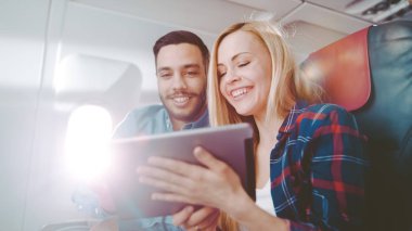 Ticari uçak bir gemide güzel genç sarışın yakışıklı İspanyol erkek kullanımı ile Tablet bilgisayar ve gülümseme. Uçak pencereden güneş parlıyor.