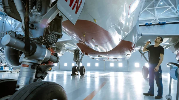 In a Hangar Aircraft Maintenance Engineer / Technician / Mechanic Visually Inspects Airplane 's Chassis and Body / Fuselage walking Under It . — стоковое фото