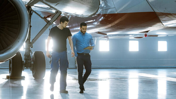 In a Hangar Aircraft Maintenance Engineer Shows  Technical Data on Tablet Computer to Airplane Technician. They Stand Near Clean Brand New Plane.