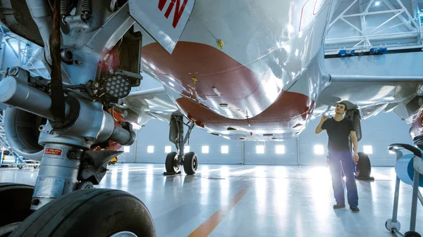 Em um Engenheiro de Manutenção de Aeronaves Hangar / Técnico / Mecânico Inspeciona Visualmente o Chassi e o Corpo / Fuselagem do Avião andando sob ele . — Fotografia de Stock