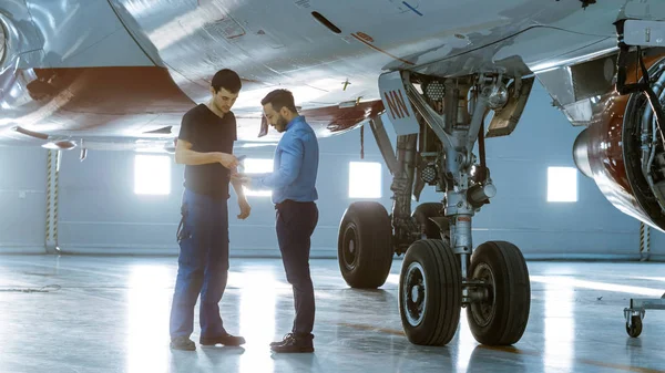 Dans un hangar, un technicien d'entretien d'aéronefs montre des données techniques sur un ordinateur tablette à un technicien d'avion. Ils se tiennent près de l'avion neuf propre . — Photo