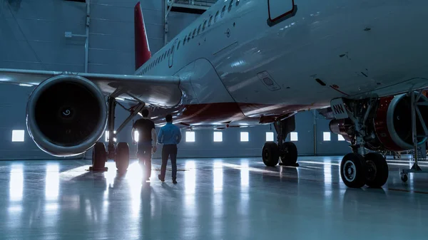 In einem Hangar zeigt ein Flugzeugwartungsingenieur dem Flugzeugtechniker technische Daten auf einem Tablet-Computer. Sie laufen neben sauberen nagelneuen Flugzeugen. — Stockfoto