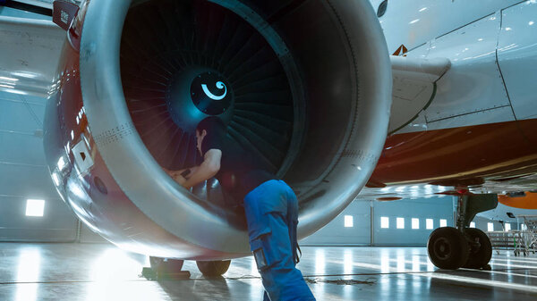 In a Hangar Aircraft Maintenance Engineer/ Technician/ Mechanic Inspects with a Flashlight Airplane's Jet Engine.