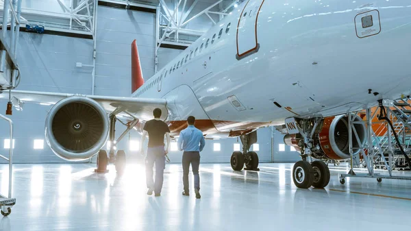 Dans un hangar, un technicien d'entretien d'aéronefs montre des données techniques sur un ordinateur tablette à un technicien d'avion. Ils marchent aux côtés de l'avion neuf propre . — Photo