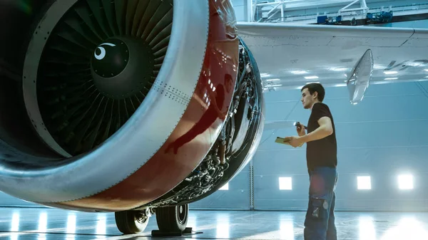 Em um Hangar Aircraft Manutenção Jovem Engenheiro / Técnico / Mecânico com Tablet Computer Inspects Airplane Jet Engine. Ele abre a escotilha do motor e examina o interior com uma lanterna elétrica . — Fotografia de Stock