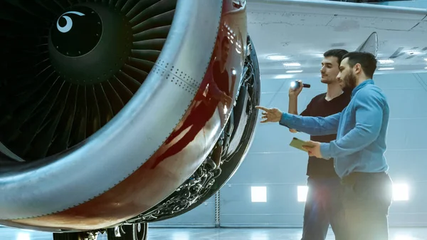 I en Hangar flygplan underhåll ingenjör visar tekniska Data på Tablet PC till flygplan tekniker diagnostisera de jetmotor genom öppen lucka. De står nära ren helt nya plan. — Stockfoto