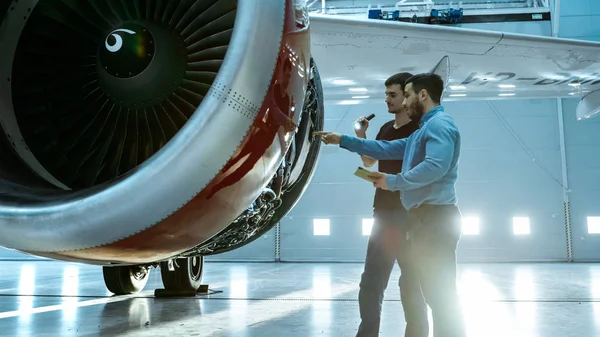 En un Hangar Aircraft Maintenance Engineer muestra datos técnicos en Tablet Computer a Airplane Technician, diagnostican el motor Jet a través de Open Hatch. Se paran cerca de nuevo avión limpio a estrenar . — Foto de Stock