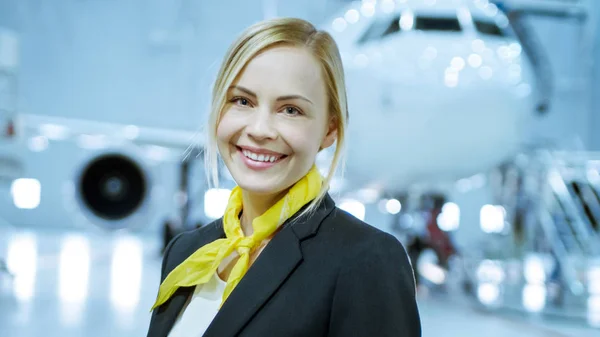 In un hangar di manutenzione aeronautica giovane bella hostess bionda / assistente di volo sorride incantevolmente sulla macchina fotografica. Sullo sfondo nuovissimo aereo è visibile . — Foto Stock