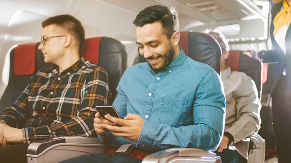 Na komercyjnych lot Młodzi Hiszpanie mężczyzna używa smartfonem, a jego sąsiad wygląda przez okno. Starszy człowiek z tyłu śpi spokojnie. — Zdjęcie stockowe