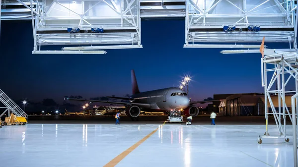 Hangar de maintenance des aéronefs où un nouvel avion est remorqué par un tracteur / remorqueur à poussée arrière sur la piste d'atterrissage. Équipage de mécaniciens, ingénieurs et conducteurs travaille activement . — Photo